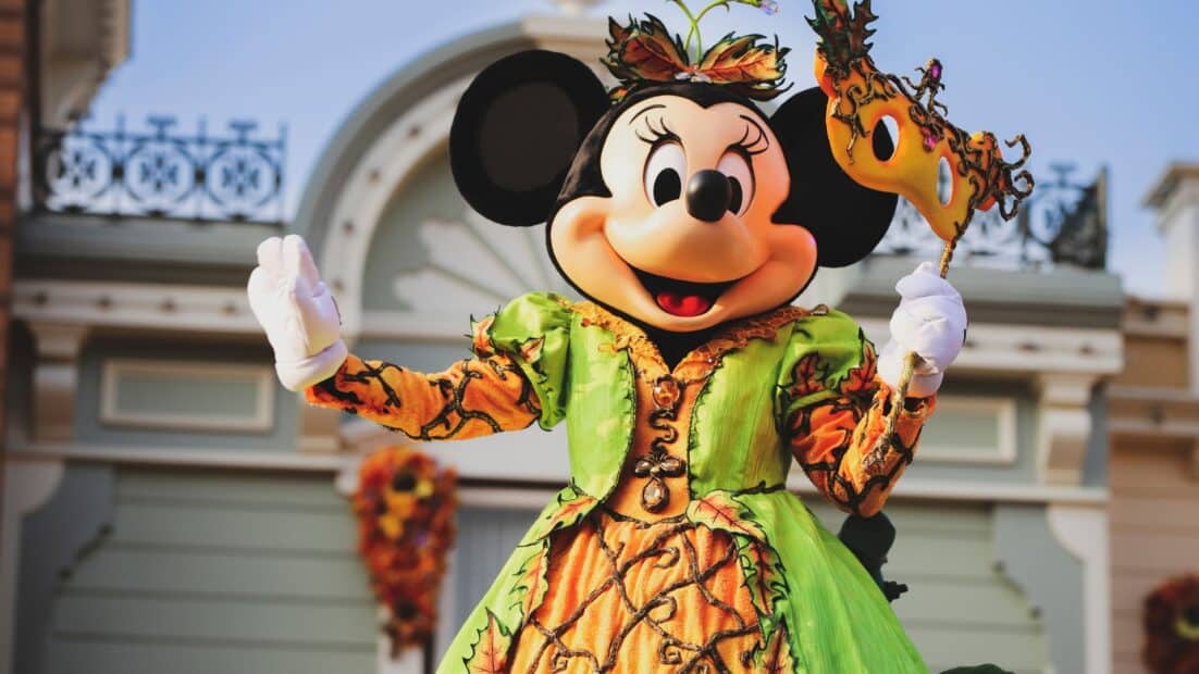 Minnie Mouse in a fall Halloween costume holding a masquerade mask during a Disney park seasonal parade